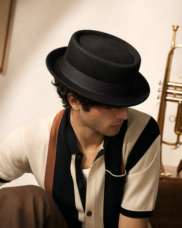 Man in a black pork pie hat and textured knit shirt beside a trumpet in warm evening light.