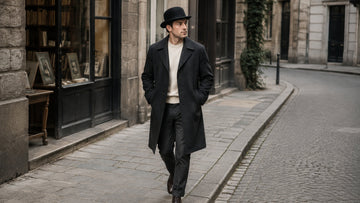 Full-length adult wearer in a black bowler hat walking through a quiet city setting in a restrained tailored outfit.
