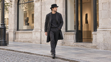 Adult walking outside a quiet stone-fronted gallery wearing a black rounded bowler hat, dark overcoat, knitwear and straight trousers.