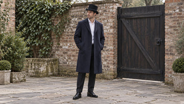 Full-body outdoor outfit with a black top hat, deep navy overcoat, pale shirt and dark trousers beside a brick wall.