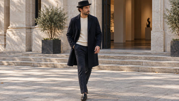 Modern fictional adult wearing a black pork pie hat with a navy overcoat, knitwear and straight trousers outside a quiet stone-fronted gallery.