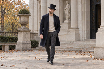 Full-body adult man in a black top hat, deep navy overcoat, oatmeal knit and charcoal trousers outside a quiet stone gallery.